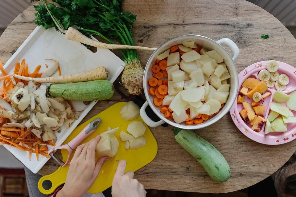 Кухонный стол с нарезанными овощами, кастрюлей и руками, чистящими картофель